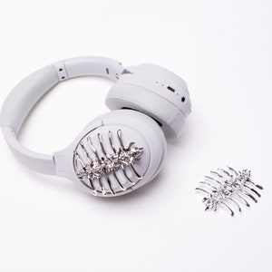 May include: White wireless headphones with a silver decorative comb attached to the earcup. A second silver comb is on the white surface.
