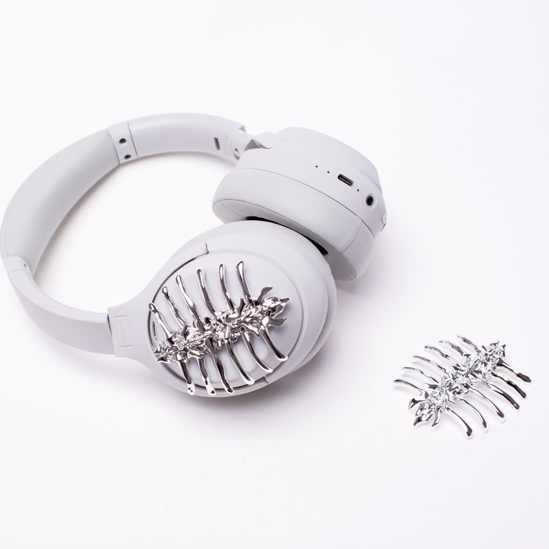 May include: White wireless headphones with a silver decorative comb attached to the earcup. A second silver comb is on the white surface.
