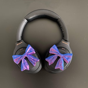 May include: Black over-ear headphones with a blue and purple iridescent bow on each earcup. The headphones have the word "SONY" printed on each earcup.