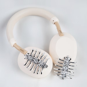 May include: Cream-colored Sony headphones with silver decorative accents. The earcups feature a silver spine-like design. The headband and earcups are a soft cream color, with the brand name "SONY" printed on the arms.