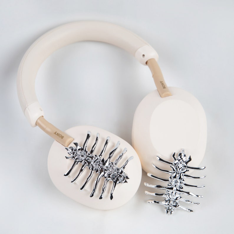 May include: Cream-colored Sony headphones with silver decorative accents. The earcups feature a silver spine-like design. The headband and earcups are a soft cream color, with the brand name "SONY" printed on the arms.