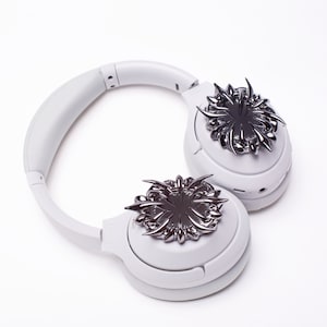 May include: White wireless headphones with black metal accents on each earcup. The accents are in the shape of a stylized starburst.