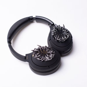 May include: Black over-ear headphones with a silver decorative design on each earcup. The design is intricate and resembles a stylized flower.