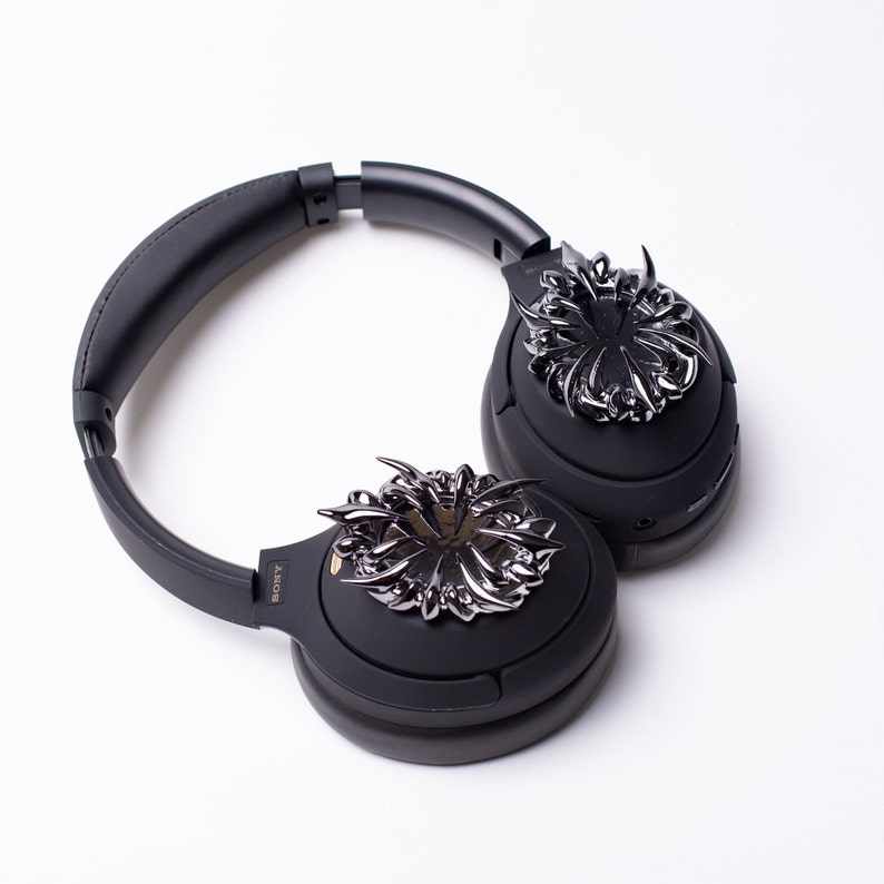 May include: Black over-ear headphones with a silver decorative design on each earcup. The design is intricate and resembles a stylized flower.