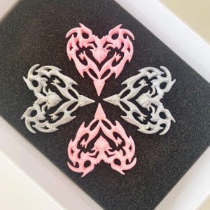May include: Four heart-shaped charms, two are pink and two are gray. Each charm has a detailed, intricate design with a pointed tip at the bottom.