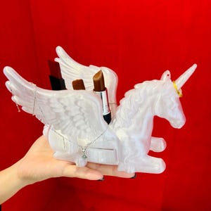 Puede incluir: Un organizador de maquillaje blanco nacarado con forma de unicornio con alas. El organizador contiene tres barras de labios. Un collar de plata con un colgante cuelga de las alas. El fondo es rojo liso.