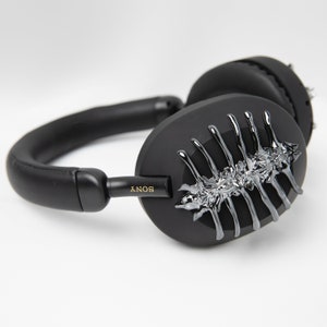 May include: Black over-ear headphones with a silver, textured, skeletal design on one earcup. The headphones have a black headband and earcups with the word "SONY" printed on the side.