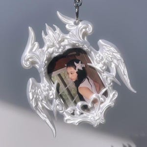 May include: A white, ornate pendant featuring a framed image of a person. The frame has a decorative, swirling design with wing-like elements. The pendant hangs from a silver chain, creating a delicate and ethereal aesthetic.