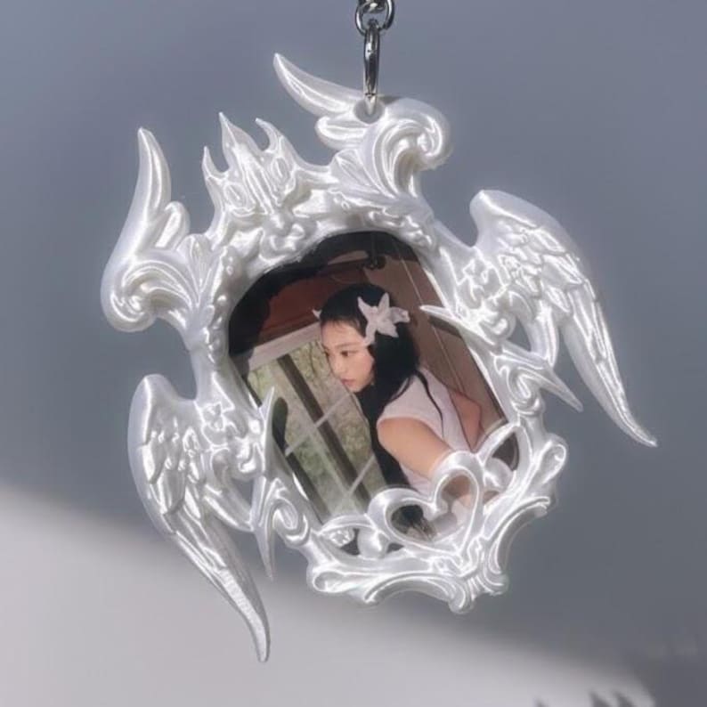 May include: A white, ornate pendant featuring a framed image of a person. The frame has a decorative, swirling design with wing-like elements. The pendant hangs from a silver chain, creating a delicate and ethereal aesthetic.