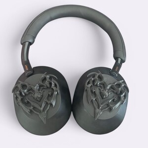May include: Black over-ear headphones with a decorative heart-shaped design on each earcup. The headphones have a black headband and the word "SONY" is printed in gold on each earcup.