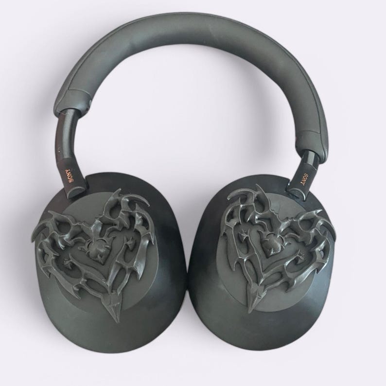 May include: Black over-ear headphones with a decorative heart-shaped design on each earcup. The headphones have a black headband and the word "SONY" is printed in gold on each earcup.
