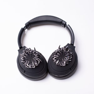 May include: Black wireless headphones with a silver, ornate design on each earcup. The design features a stylized, geometric pattern with sharp points.