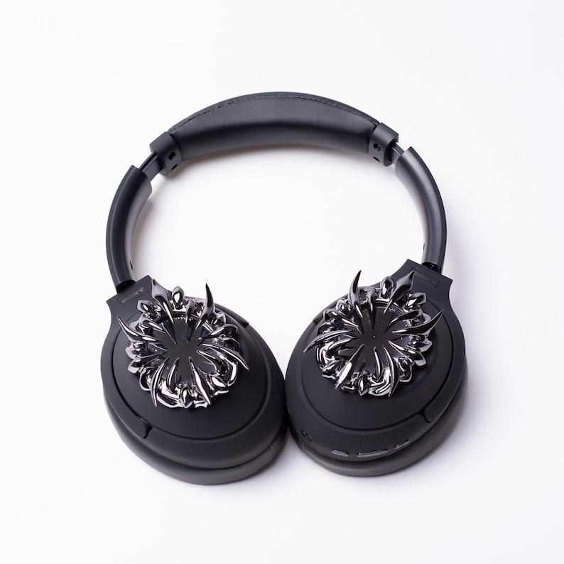 May include: Black wireless headphones with a silver, ornate design on each earcup. The design features a stylized, geometric pattern with sharp points.