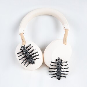 May include: Cream-colored Sony headphones with black, spine-like embellishments on the earcups. The headband and earcups are a soft cream color, with tan-colored arms. The Sony logo is visible on the arms.