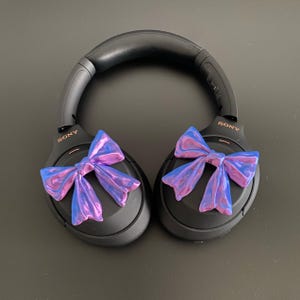 May include: Black over-ear headphones with a pink and blue iridescent bow on each earcup. The headphones have the word "SONY" printed on the side.