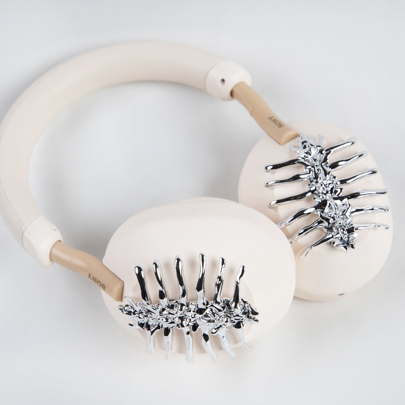 May include: Cream-colored headphones with silver-colored spine-like embellishments on the earcups. The headband and earcups are a soft cream color, with the brand name "SONY" printed on the arms.