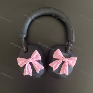 May include: Black over-ear headphones with pink bows on each earcup. The headphones have a black headband and the earcups are round. The text "SONY" is visible on the earcups.