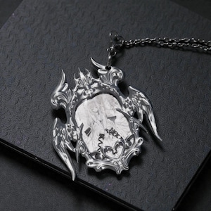 May include: A silver-colored pendant necklace with a detailed, ornate frame. The pendant features a black and white illustration within the frame, and is suspended from a matching chain. The design has a gothic or fantasy theme.