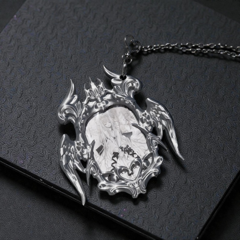May include: A silver-colored pendant necklace with a detailed, ornate frame. The pendant features a black and white illustration within the frame, and is suspended from a matching chain. The design has a gothic or fantasy theme.