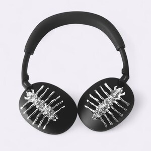 May include: Black over-ear headphones with silver-colored decorative accents. The earcups are oval-shaped and feature a silver-colored design. The headband and earcups are black. The headphones are on a white background.