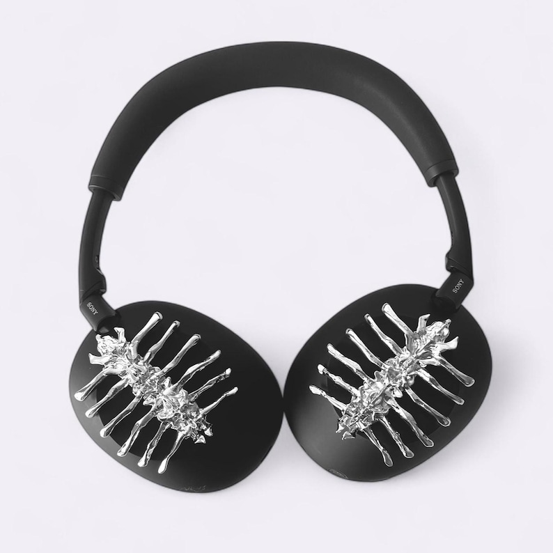 May include: Black over-ear headphones with silver-colored decorative accents. The earcups are oval-shaped and feature a silver-colored design. The headband and earcups are black. The headphones are on a white background.