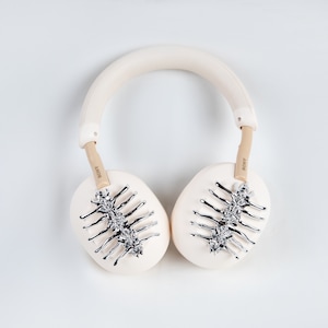 May include: Cream-colored over-ear headphones with silver decorative accents. The earcups feature a silver spine-like design. The headband and earcups are a soft cream color, with the brand name "SONY" visible on the arms.