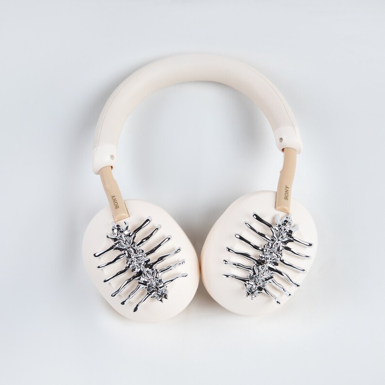 May include: Cream-colored over-ear headphones with silver decorative accents. The earcups feature a silver spine-like design. The headband and earcups are a soft cream color, with the brand name "SONY" visible on the arms.