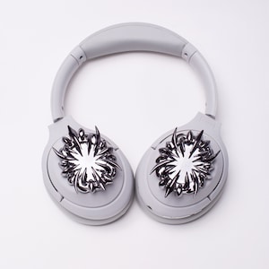 May include: White headphones with silver decorative accents on each earcup. The accents are stylized and resemble a flower or starburst.