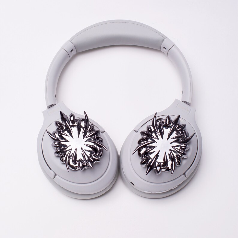 May include: White headphones with silver decorative accents on each earcup. The accents are stylized and resemble a flower or starburst.