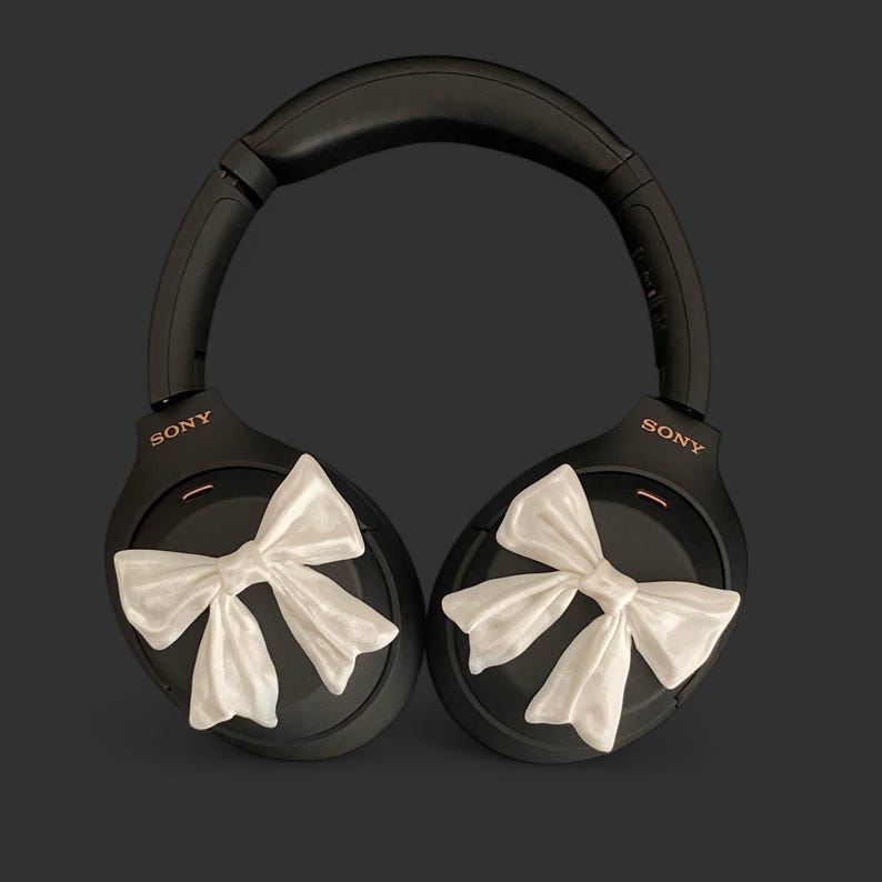 K&ouml;nnte beinhalten: Schwarze Over-Ear-Kopfh&ouml;rer mit wei&szlig;en Schleifen an jedem Ohrpolster. Die Kopfh&ouml;rer haben den Schriftzug "SONY" in Ros&eacute;gold auf jedem Ohrpolster.