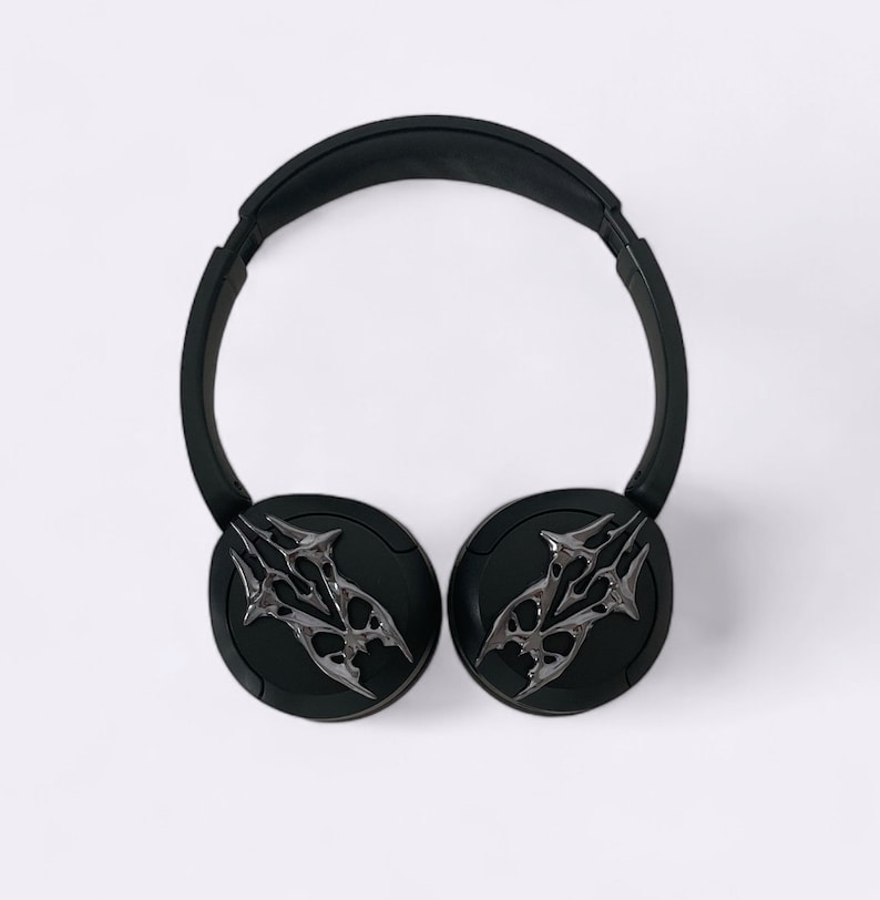 Puede incluir: Auriculares inal&aacute;mbricos negros con un dise&ntilde;o plateado en las almohadillas. El dise&ntilde;o presenta un patr&oacute;n de llama estilizado.