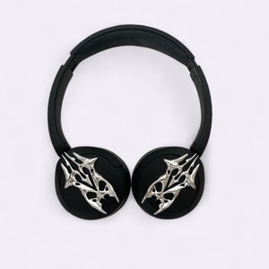 Puede incluir: Auriculares inal&aacute;mbricos negros con detalles decorativos plateados en las almohadillas. Los detalles son estilizados y se asemejan a un par de garras.