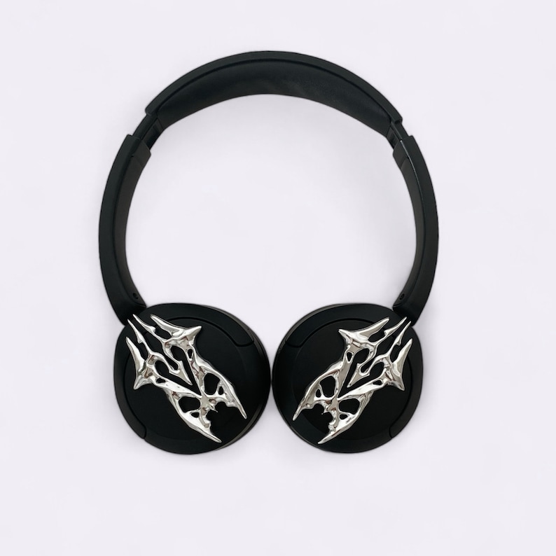 Puede incluir: Auriculares inal&aacute;mbricos negros con detalles decorativos plateados en las almohadillas. Los detalles son estilizados y se asemejan a un par de garras.