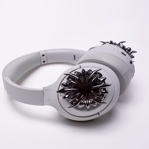 May include: White wireless headphones with a silver, spiky design on one earcup. The headphones have a black logo on the side.