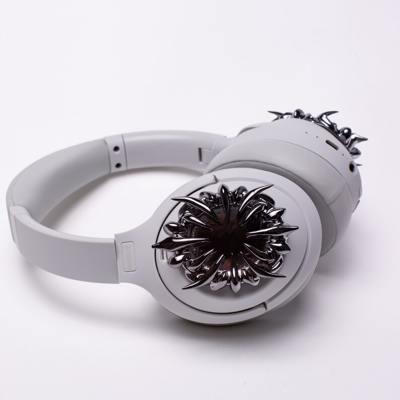 May include: White wireless headphones with a silver, spiky design on one earcup. The headphones have a black logo on the side.