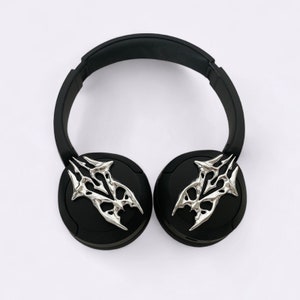 Puede incluir: Auriculares negros con detalles decorativos plateados en las almohadillas. Los detalles tienen un dise&ntilde;o estilizado y abstracto.