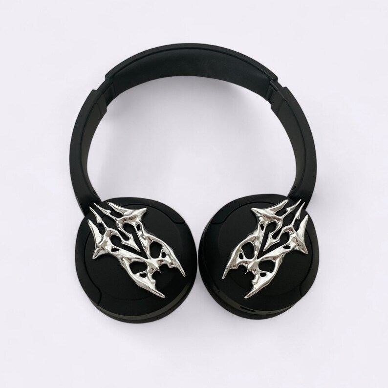 Puede incluir: Auriculares negros con detalles decorativos plateados en las almohadillas. Los detalles tienen un dise&ntilde;o estilizado y abstracto.