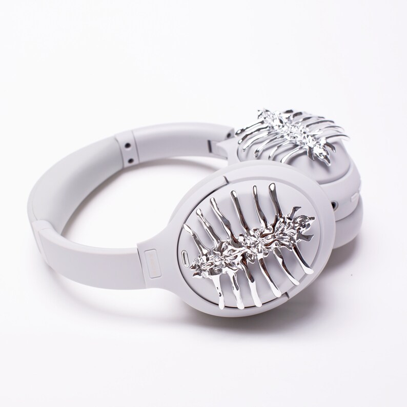 May include: Gray over-ear headphones with a silver decorative design on each earcup. The design resembles a stylized skeletal structure with intricate details.