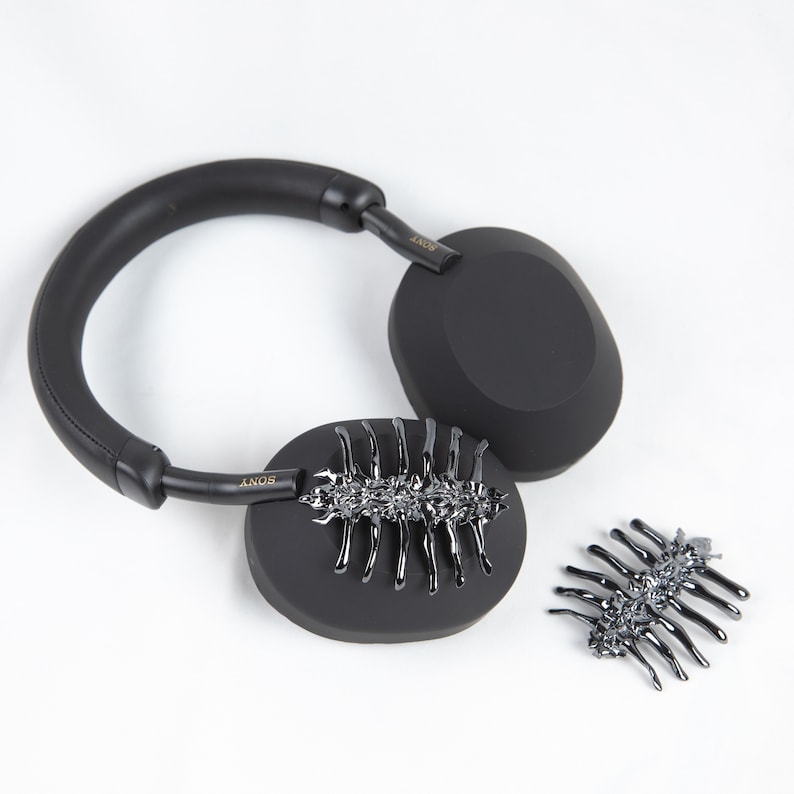 May include: Black over-ear headphones with a silver decorative element on each earcup. The decorative element resembles a stylised insect.