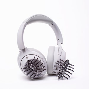 May include: A pair of white wireless headphones with a decorative black metal hair clip on each earcup.