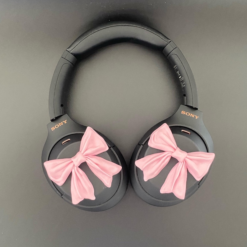 K&ouml;nnte beinhalten: Schwarze Over-Ear-Kopfh&ouml;rer mit rosa Schleifen an jedem Ohrpolster. Die Kopfh&ouml;rer haben den Schriftzug "SONY" in goldener Schrift auf jedem Ohrpolster.