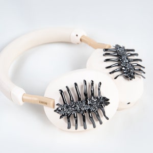 May include: Cream-colored over-ear headphones with black, spine-like embellishments on the earcups. The headband and earcups are a soft, neutral tone, with the brand name "SONY" visible on the side. The design features a unique, dark, glossy accent.