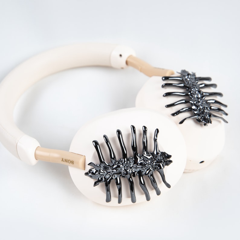 May include: Cream-colored over-ear headphones with black, spine-like embellishments on the earcups. The headband and earcups are a soft, neutral tone, with the brand name "SONY" visible on the side. The design features a unique, dark, glossy accent.