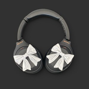 K&ouml;nnte beinhalten: Schwarze Over-Ear-Kopfh&ouml;rer mit wei&szlig;en Schleifen an jedem Ohrpolster. Die Kopfh&ouml;rer tragen den Schriftzug "SONY" auf jedem Ohrpolster.