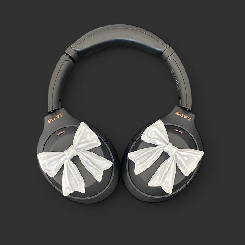 K&ouml;nnte beinhalten: Schwarze Over-Ear-Kopfh&ouml;rer mit wei&szlig;en Schleifen an jedem Ohrpolster. Die Kopfh&ouml;rer tragen den Schriftzug "SONY" auf jedem Ohrpolster.