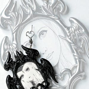 May include: A black framed pendant with a gothic design, featuring a portrait of a woman. The pendant is suspended from a silver chain with a heart-shaped clasp. A larger, silver-colored frame with an angel-wing design is visible in the background.