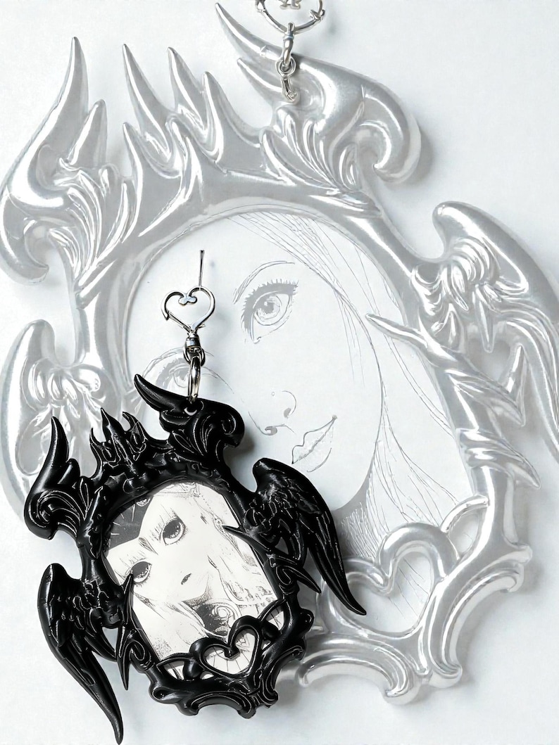 May include: A black framed pendant with a gothic design, featuring a portrait of a woman. The pendant is suspended from a silver chain with a heart-shaped clasp. A larger, silver-colored frame with an angel-wing design is visible in the background.