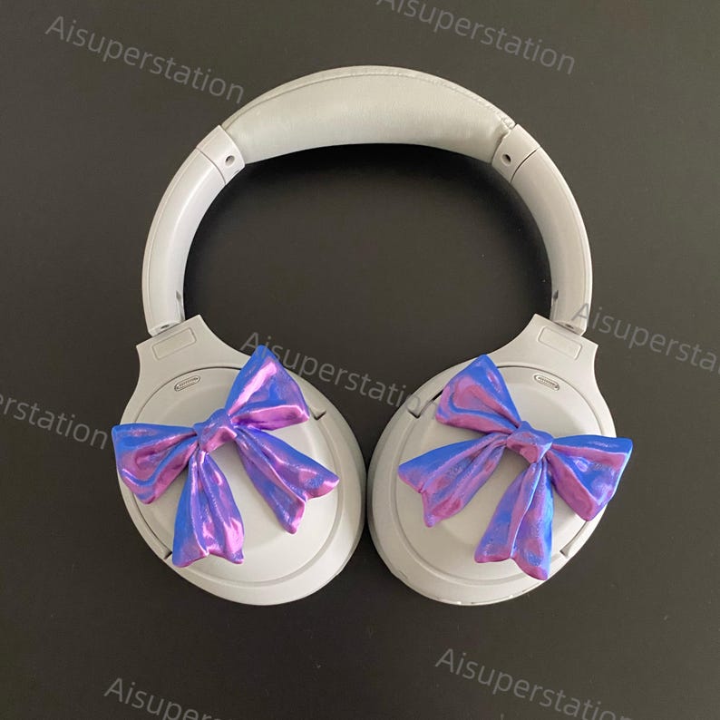 May include: A pair of white noise-cancelling headphones with iridescent purple and blue bows attached to the earcups.