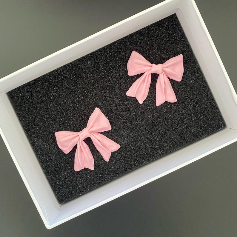 K&ouml;nnte beinhalten: Zwei rosa Stoffschleifen in einer wei&szlig;en Schachtel mit schwarzem Schaumstoff-Innenleben.