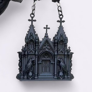 May include: A black gothic cathedral-shaped pendant with intricate architectural details. The pendant features two towers, a central cross, and a detailed facade with a pair of ravens perched on skulls. The pendant is suspended by a chain.